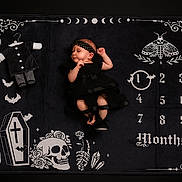 Lilith is registered to the contest to win money with this photo: baby, infant, black_dress, headband, milestone_blanket, skull, coffin, bats, crystals, months, one_month, lying_down, peaceful, gothic, dark_theme, portrait, cute, newborn, decorative_blanket, baby_milestone