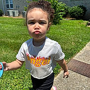 Kinley joined the competition — help win amazing prizes! child, toddler, curly_hair, white_shirt, angel_text, flame_design, black_shorts, pink_shoes, sidewalk, grass, bushes, building, windows, sunny, outdoor, pouting, walking, holding_object, shadow, daylight