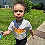 child, toddler, curly_hair, white_shirt, angel_text, flame_design, black_shorts, pink_shoes, sidewalk, grass, bushes, building, windows, sunny, outdoor, pouting, walking, holding_object, shadow, daylight