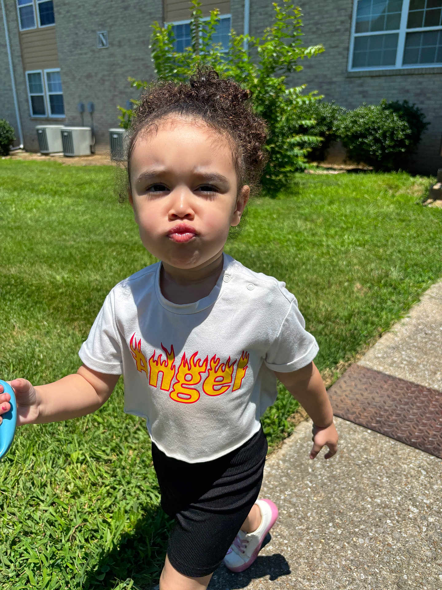 Kinley joined the competition — help win amazing prizes! child, toddler, curly_hair, white_shirt, angel_text, flame_design, black_shorts, pink_shoes, sidewalk, grass, bushes, building, windows, sunny, outdoor, pouting, walking, holding_object, shadow, daylight