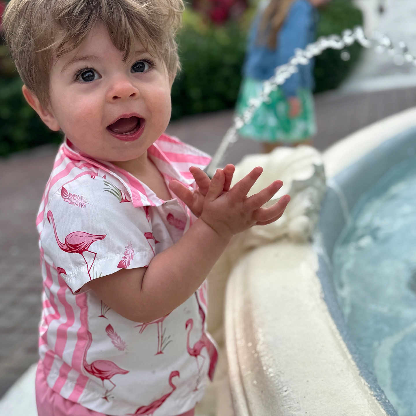 Callaghan is registered to the contest to win money with this photo: blond_hair, blurred_background, casual_clothing, child, clapping, curly_hair, daylight, flamingo_shirt, fountain, fun, garden, happy, outdoor, person, pink_pants, playful, smiling, toddler, water, young_child
