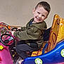 child, smiling, happy, indoors, toy_car, ride, steering_wheel, colorful, seat, footwear, jacket, pants, play, fun, young, person, face, seatbelt, floor, wall