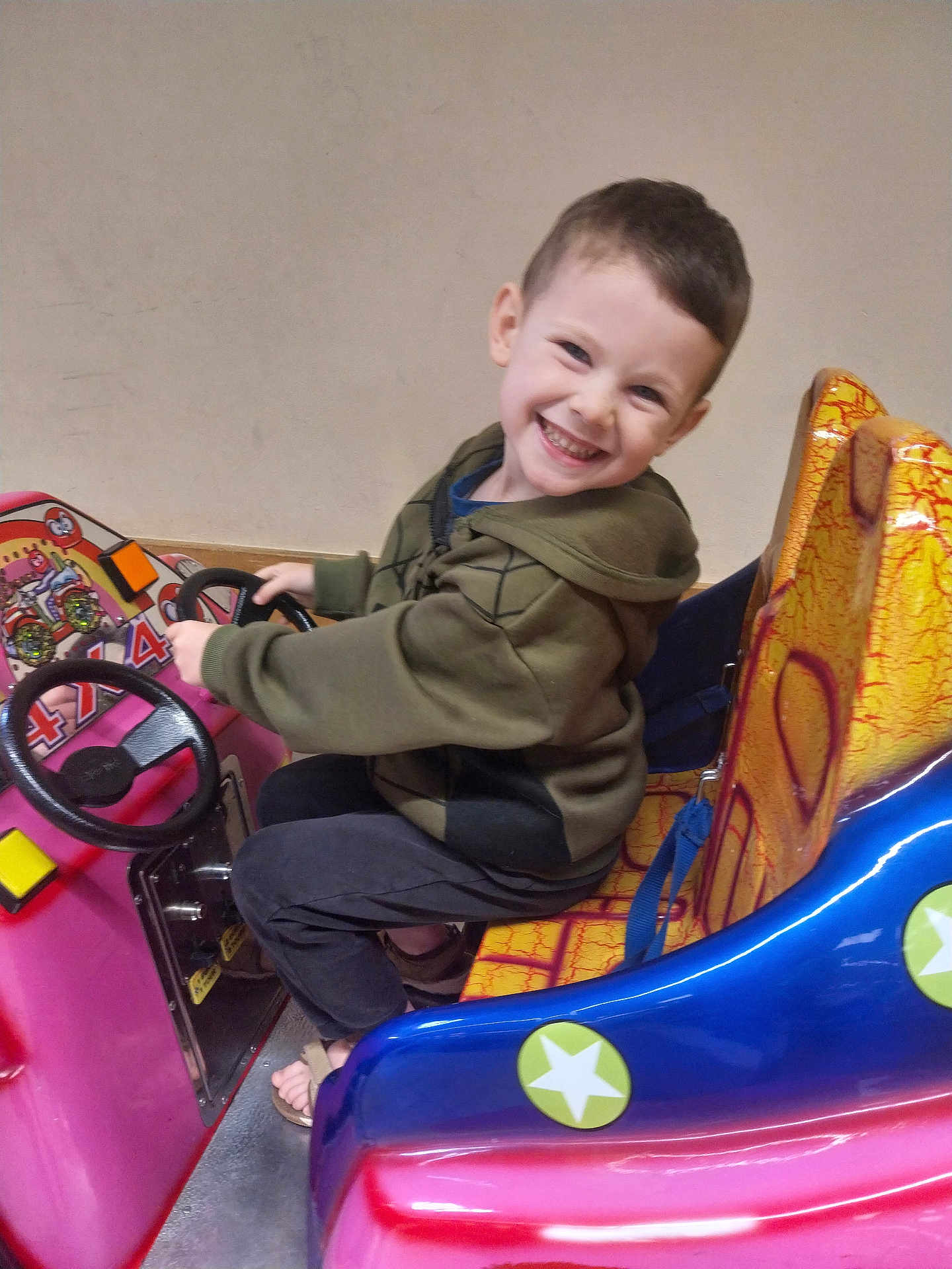 Alex a rejoint le concours — aidez-le/la à gagner de superbes lots ! child, smiling, happy, indoors, toy_car, ride, steering_wheel, colorful, seat, footwear, jacket, pants, play, fun, young, person, face, seatbelt, floor, wall