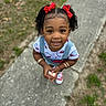 child, girl, smiling, curly_hair, red_bows, white_tshirt, denim_overalls, bracelets, sneakers, outdoor, sidewalk, grass, holding_object, portrait, cute, happy, young, fashion, accessories, standing