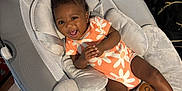 Paris is registered to the contest to win money with this photo: baby, infant, smiling, baby_seat, onesie, orange_clothing, floral_pattern, happy, child, indoor, fabric, cushion, socks, person, young_child, cute, relaxed, soft_texture, home, baby_accessory