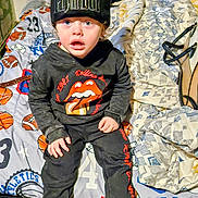 Donnavee is registered to the contest to win money with this photo: child, toddler, beanie, hat, playboy_logo, surprised_expression, sitting, bed, blanket, patterned_bedding, boots, hoodie, rolling_stones_logo, black_clothing, indoor, bedroom, curly_hair, purse_strap, eye_contact, portrait