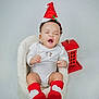 baby, infant, santa_hat, red_socks, yawning, white_onesie, cozy_seat, decorative_house, holiday_theme, cute, indoor, child, portrait, festive, smiling, soft_texture, holiday_decor, adorable, sitting, young_child