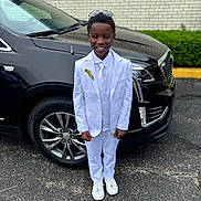 Antwon is registered to the contest to win money with this photo: child, boy, smiling, white_suit, tie, sunglasses, car, black_suv, outdoor, pavement, ribbon, formal_wear, shoes, standing, happy, person, event, vehicle, urban, daytime