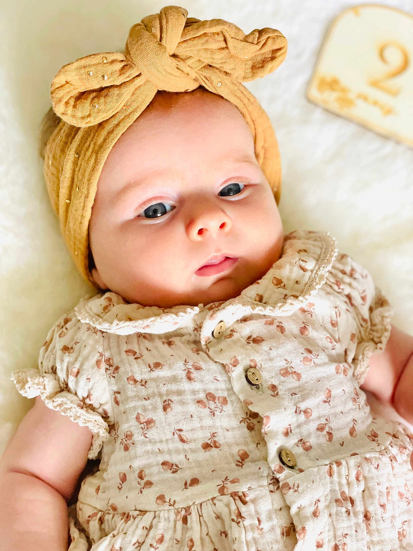 Agathe participe au concours pour gagner de l'argent avec cette photo : baby, infant, headband, yellow_headband, bow, dress, floral_pattern, buttoned_dress, soft_blanket, wooden_block, number_two, milestone, portrait, closeup, cute, child, skin, face, lying_down, adorable
