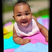 Harlow is registered to the contest to win money with this photo: baby, smiling, outdoor, blanket, colorful, child, happy, face, skin, toddler, sunlight, cute, portrait, infant, clothing, young, baby_bib, lying_down, cheerful, nature
