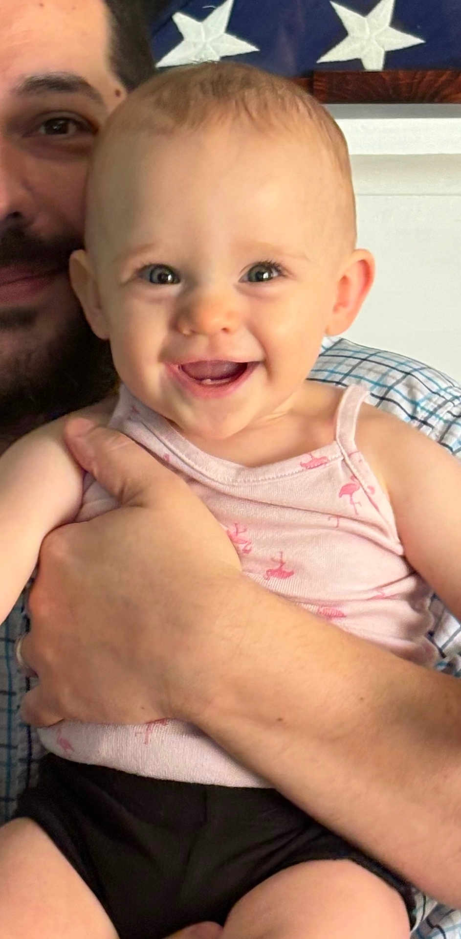 Audri is registered to the contest to win money with this photo: baby, smiling, adult, holding, happy, face, blue_eyes, tank_top, indoor, person, hand, closeup, portrait, skin, cheerful, expression, casual_clothing, background, flag, family