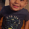 child, boy, toddler, portrait, smiling, happy, indoors, standing, t_shirt, graphic_shirt, short_hair, blond_hair, face, eyes, mouth, wooden_floor, furniture, home, photography, close_up