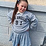 child, girl, pose, grey_sweatshirt, skirt, hair_bows, black_shoes, socks, outdoor, sidewalk, wall, confident, casual_clothing, portrait, young_person, fashion, street, smile, hands_on_hips, daylight