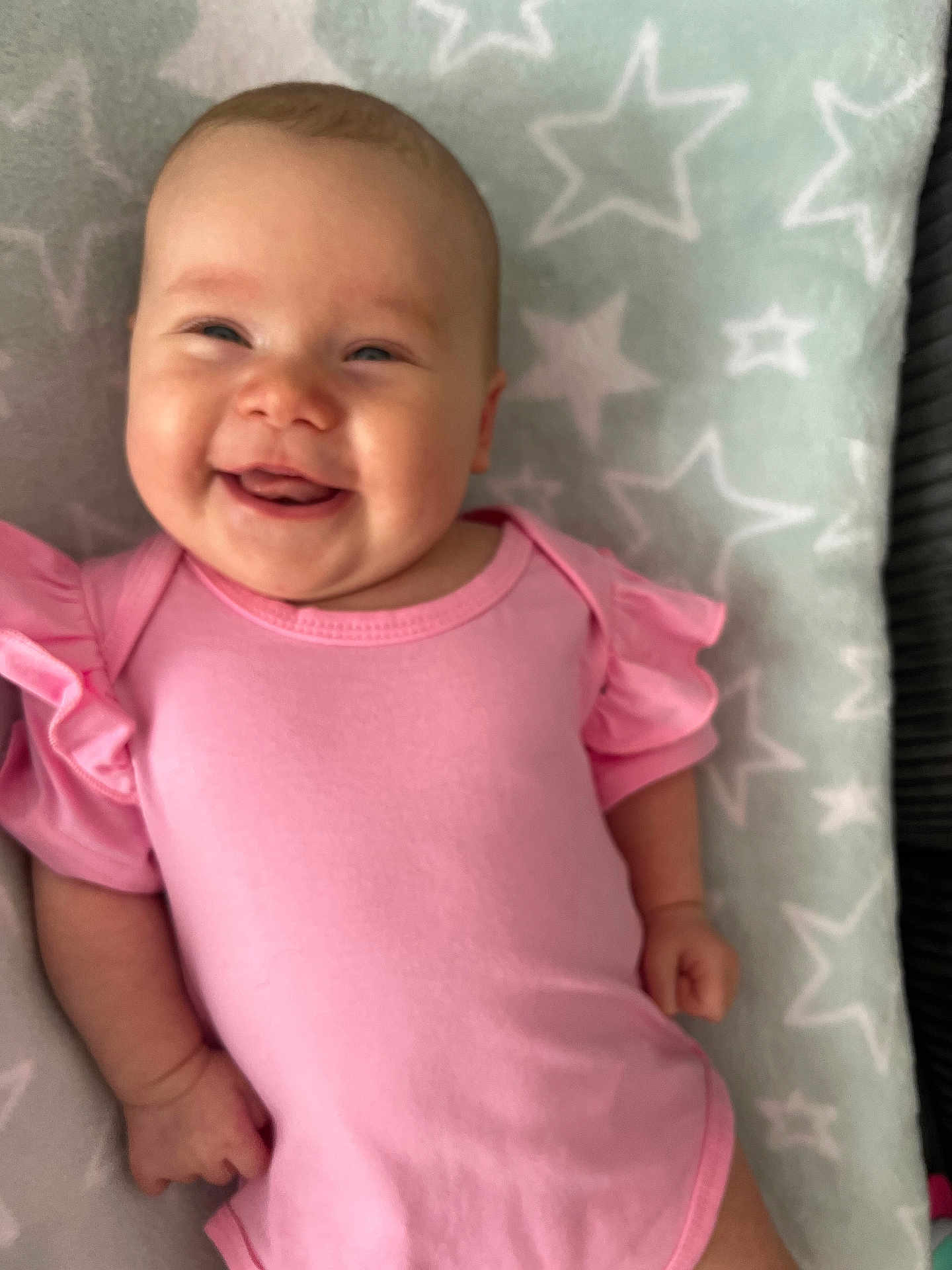Mila is registered to the contest to win money with this photo: baby, smiling, infant, pink_clothing, blanket, stars_pattern, happy, face, lying_down, cute, child, soft_texture, portrait, skin, hand, head, cheeks, person, toddler, indoors