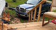 Outlaw is registered to the contest to win money with this photo: dog, porch, wooden_floor, pickup_truck, grass, trees, chair, collar, outdoor, house, backyard, vehicle, nature, daylight, relaxed, canine, leash, fence, rural, quiet