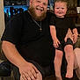 Kyle joined the competition — help win amazing prizes! man, child, toddler, beard, smile, couch, indoor, furniture, black_clothing, happy, family, person, living_room, cross_necklace, skin, hand, legs, barefoot, seated, casual