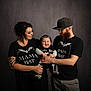 Arthur is registered to the contest to win money with this photo: baby, man, woman, family, portrait, smile, black_clothing, tattoo, beard, hat, glasses, indoor, studio, dark_background, holding, happy, tshirt, matching_outfits, child, parent