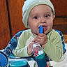 Noah is registered to the contest to win money with this photo: baby, infant, child, sitting, activity_center, toy, teething, blue_toy, green_clothing, hat, smiling, face, portrait, indoors, wooden_background, high_chair, colorful, hands, playtime, close_up