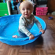Noah joined the competition — help win amazing prizes! baby, child, infant, kiddie_pool, blue_pool, kneeling, sitting, curious, surprised, open_mouth, indoors, playroom, toys, toy_storage, plastic_baskets, wood_floor, long_sleeves, pants, portrait, timestamp