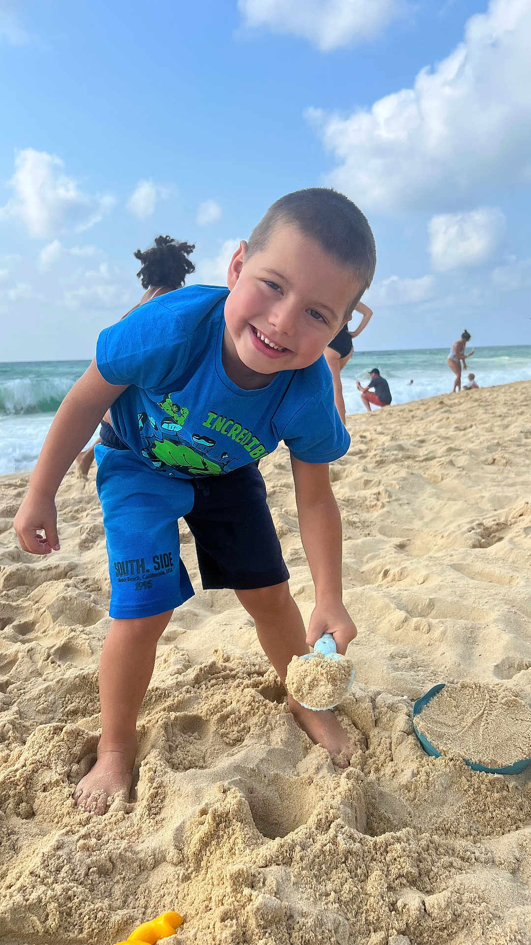 Leo participe au concours pour gagner de l'argent avec cette photo : child, boy, beach, sand, shovel, ocean, waves, blue_sky, clouds, playing, summer, vacation, smiling, shorts, tshirt, barefoot, people, sunny, outdoor, fun