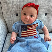 Amelia is registered to the contest to win money with this photo: baby, infant, child, red_headband, blue_dress, american_flag, knitted_clothing, armchair, textured_fabric, cute, smiling, portrait, seated, indoors, furniture, person, head, face, hand, skin