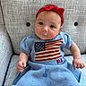 baby, infant, child, red_headband, blue_dress, american_flag, knitted_clothing, armchair, textured_fabric, cute, smiling, portrait, seated, indoors, furniture, person, head, face, hand, skin