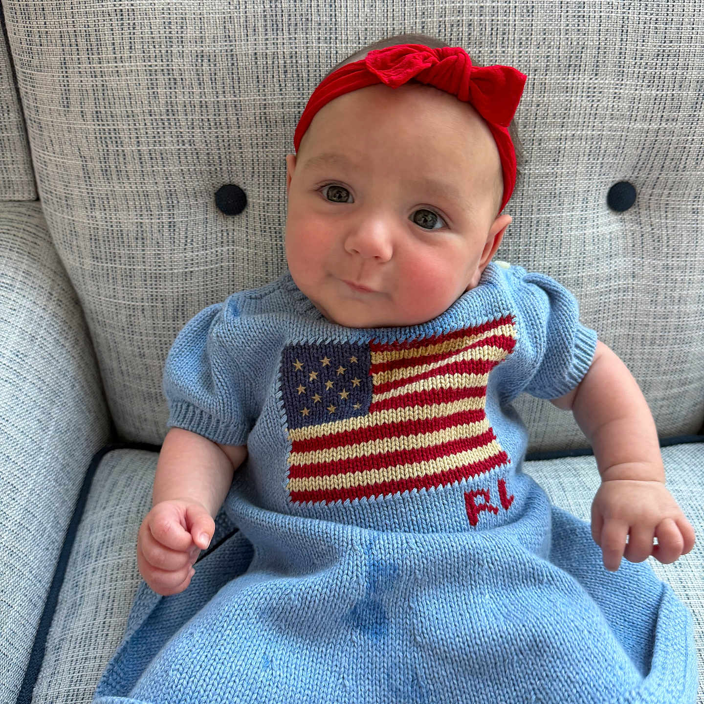 Amelia is registered to the contest to win money with this photo: baby, infant, child, red_headband, blue_dress, american_flag, knitted_clothing, armchair, textured_fabric, cute, smiling, portrait, seated, indoors, furniture, person, head, face, hand, skin