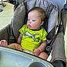 baby, child, stroller, seat, green_shirt, footwear, picnic_table, outdoor, infant, person, face, head, clothing, foot, expression, curious, summer, daylight, fabric, comfort