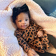 Yora joined the competition — help win amazing prizes! baby, child, infant, black_bow, curly_hair, brown_dress, heart_pattern, pink_blanket, soft_texture, smiling, happy, cute, indoors, cozy, couch, hand, face, person, portrait, clothing