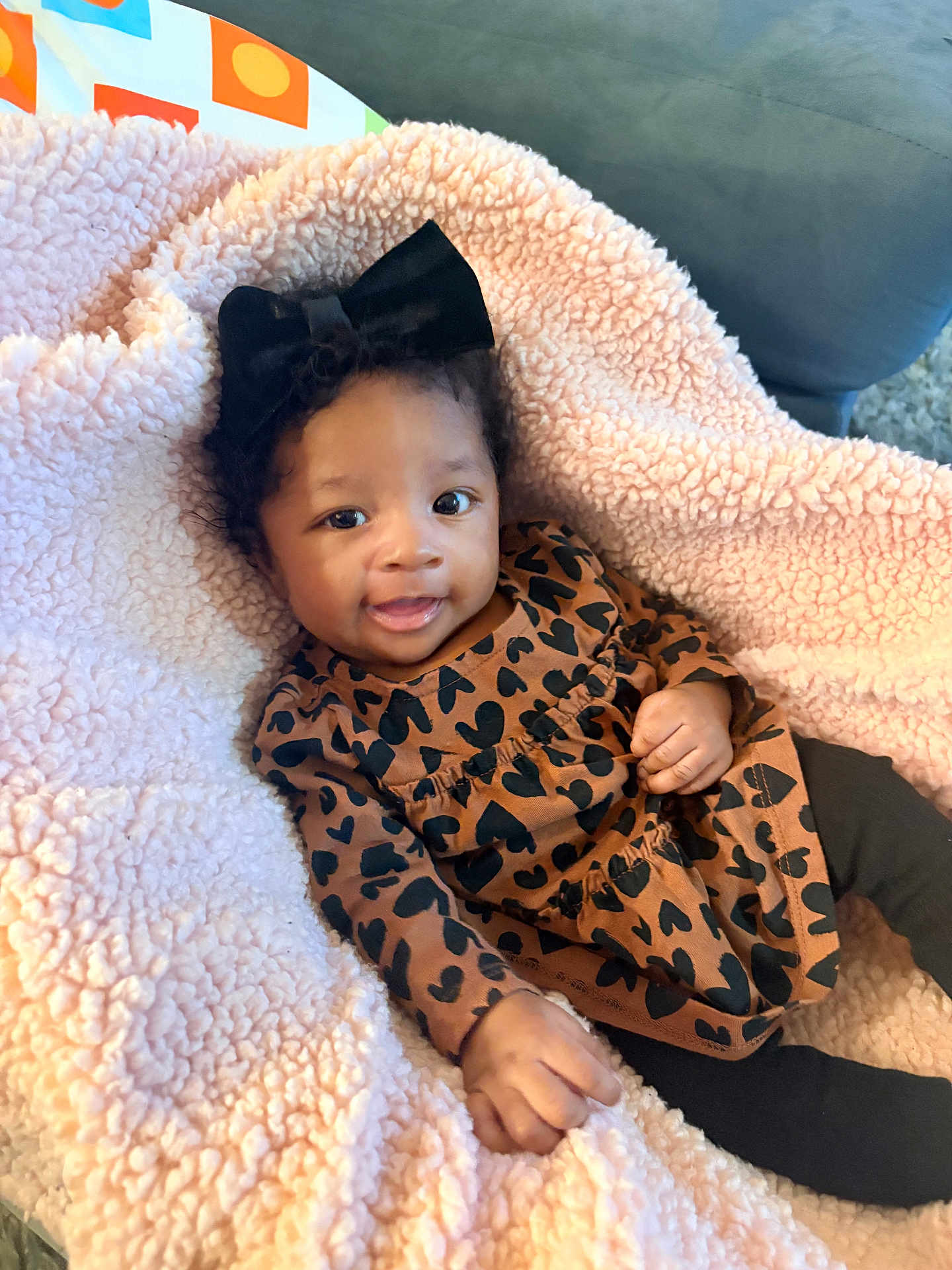 Yora joined the competition — help win amazing prizes! baby, child, infant, black_bow, curly_hair, brown_dress, heart_pattern, pink_blanket, soft_texture, smiling, happy, cute, indoors, cozy, couch, hand, face, person, portrait, clothing