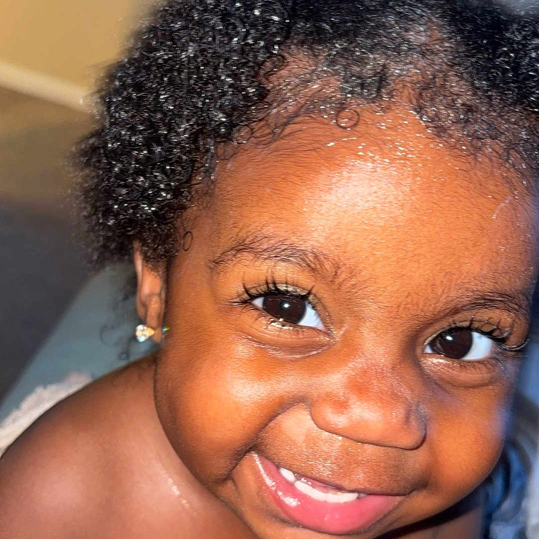 Kind is registered to the contest to win money with this photo: baby, child, closeup, curly_hair, cute, earrings, expression, eyes, face, happy, indoors, joyful, light, person, portrait, skin, smiling, toddler, towel, young_child