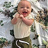Tanner is registered to the contest to win money with this photo: baby, infant, child, smiling, happy, portrait, lying_down, indoors, blanket, patterned_blanket, pinecones, plants, clothing, cream_shirt, green_pants, hands, face, eyes, mouth, photography