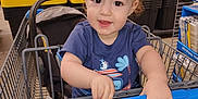 Andrew is registered to the contest to win money with this photo: aisle, baby, black_storage_bins, blue_clothing, casual_wear, child, curly_hair, footwear_absent, happy, holding, indoor, metal_cart, person, retail, seat, shopping_cart, smiling, store, toddler, yellow_storage_bins