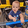 toddler, child, shopping_cart, store, aisle, blue_clothing, smiling, holding, yellow_storage_bins, black_storage_bins, indoor, baby, person, retail, curly_hair, happy, seat, metal_cart, footwear_absent, casual_wear