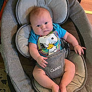 Axel is registered to the contest to win money with this photo: baby, infant, baby_rocker, snoopy, tshirt, blue_clothing, indoor, carpet, wood_paneling, baby_seat, child, person, face, legs, feet, expression, furniture, floor, home, cute