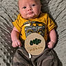 baby, infant, one_month, yellow_shirt, camo_pants, gray_blanket, wooden_sign, milestone, portrait, cute, child, lying_down, socks, indoors, person, face, young_baby, smiling, curious, soft_texture