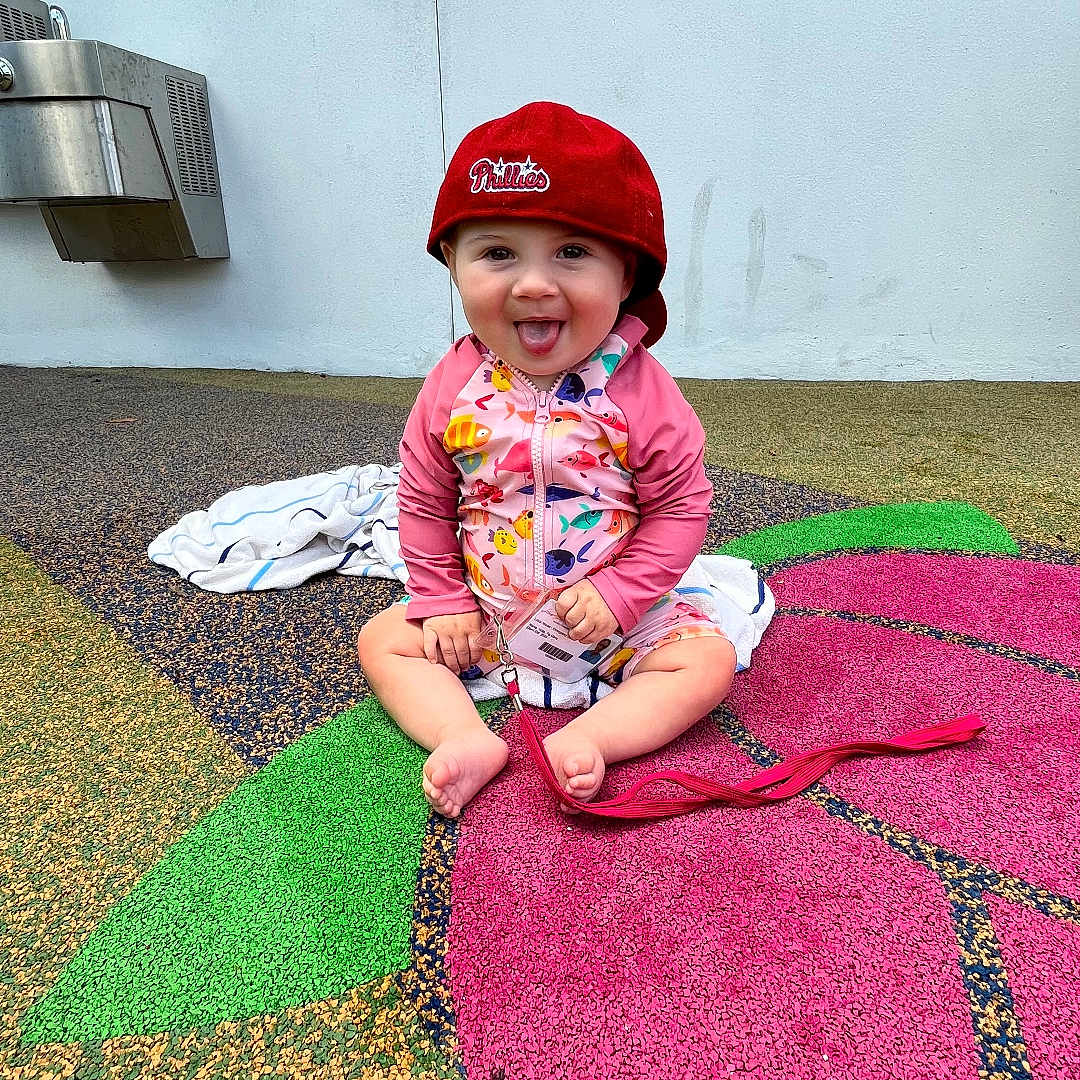 Indie Ana is registered to the contest to win money with this photo: barefoot, casual, child, colorful_clothing, floor_pattern, flower_shape, fun, hat, outdoor, person, philadelphia_phillies, playful, playground, red_cap, sitting, smiling, toddler, tongue_out, towel, young_child