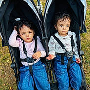 Tara is registered to the contest to win money with this photo: toddler, child, stroller, outdoor, grass, blue_pants, long_sleeve, barefoot, curly_hair, matching_outfits, seatbelt, playground, park, person, two_children, serious_expression, daylight, nature, casual_clothing, sitting