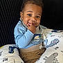 baby, child, smiling, blue_shirt, beige_pants, dinosaur_cushion, pacifier, sitting, indoor, black_background, curly_hair, playful, cute, happy, infant, portrait, cushion, clothing, person, face