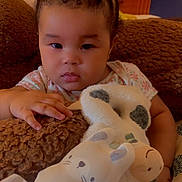 Truth is registered to the contest to win money with this photo: baby, child, plush_toy, stuffed_animal, cow, cat_toy, bow, hair_accessory, bed, pillow, cushion, wooden_headboard, indoor, infant, hand, clothing, face, expression, person, soft_toys