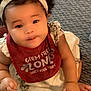 baby, child, infant, headband, bib, white_dress, carpet, sitting, cute, smiling, hand, curly_hair, indoor, young_child, person, face, looking_up, red_bib, skin, clothing