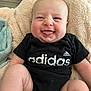 baby, infant, smiling, tongue_out, onesie, black_clothing, blanket, fluffy, indoor, cute, chubby_cheeks, happy, person, skin, head, face, sitting, comfortable, soft_texture, adidas