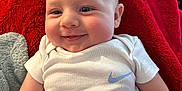 Jaxx joined the competition — help win amazing prizes! baby, infant, smile, white_onesie, nike_logo, red_blanket, plush_toy, indoor, living_room, child, cute, happy, face, skin, soft_texture, seated, person, head, cheeks, casual
