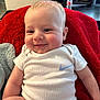 baby, infant, smile, white_onesie, nike_logo, red_blanket, plush_toy, indoor, living_room, child, cute, happy, face, skin, soft_texture, seated, person, head, cheeks, casual