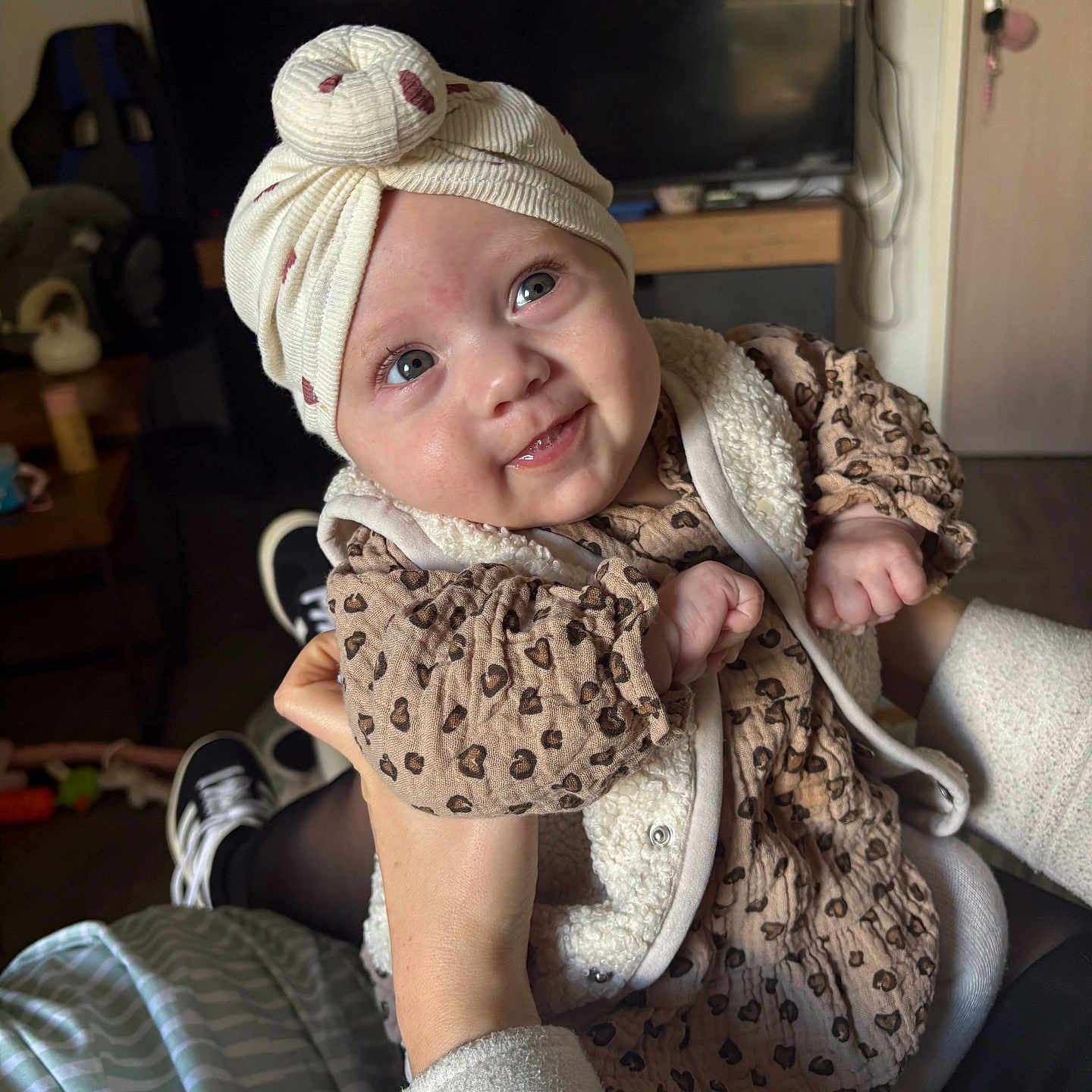 Romy a rejoint le concours — aidez-le/la à gagner de superbes lots ! baby, being_held, child, cozy, door, eyes, hands, hat, headwrap, indoor, infant, leopard_print, living_room, patterned_clothing, portrait, seated, smiling, turban, tv, vest