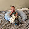 baby, smiling, plush_toy, blanket, cushion, gray, blue, clothing, sitting, indoor, happy, child, infant, person, footwear, pacifier, neutral_background, cute, portrait, cozy