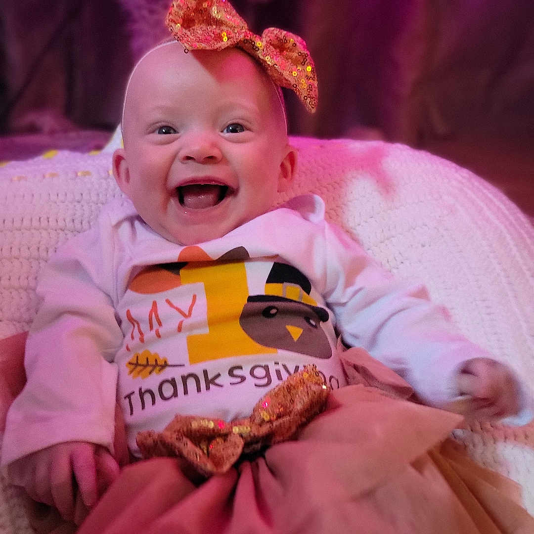 Magnolia is registered to the contest to win money with this photo: baby, smiling, thanksgiving, bow, skirt, pink_lighting, holiday, celebration, infant, cute, festive, happy, clothing, blanket, portrait, indoors, child, toddler, face, sitting