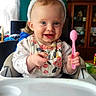 baby, bunny_ears, high_chair, pink_spoon, smiling, child, indoor, person, cute, happy, feeding_time, clothing, face, blue_eyes, table, furniture, home, cabinet, curtains, playful