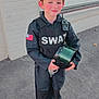 black_clothing, boy, brick_wall, child, costume, footwear, helmet, holding, outdoor, person, radio, red_cheeks, serious_expression, sidewalk, standing, swat, uniform, urban, window, young
