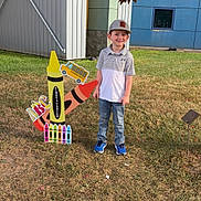 Braxton is registered to the contest to win money with this photo: boy, building, cap, casual_clothing, child, crayons, daylight, educational, grass, jeans, kindergarten, leaves, outdoor, playful, school_bus, sidewalk, sign, smiling, sneakers, tree